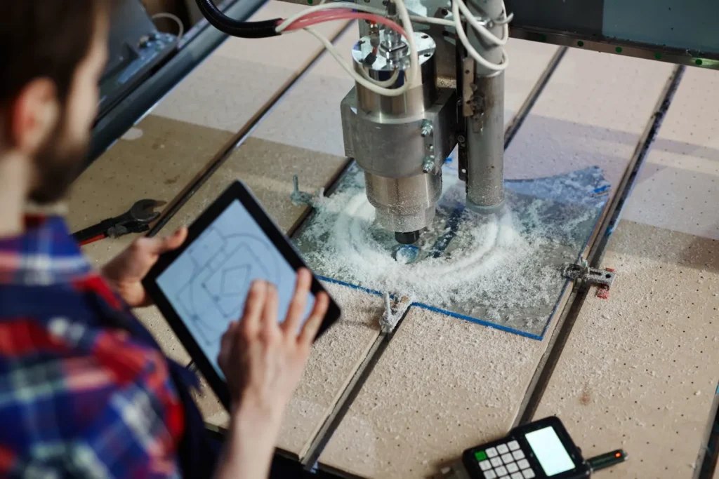 A machinist at a CNC router checking the part drawing on a tablet while the spindle cuts.