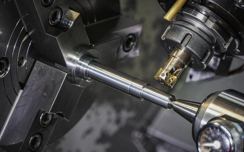 A metal shaft held in a lathe chuck with live tooling cutting it.